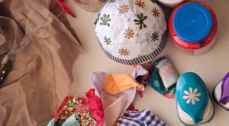 Tecidos coloridos, recortes de papéis, tesoura, grampeador e uma embalagem de ovo de Páscoa decorado com colagens. Todos os objetos apoiados em uma superfície lisa branca